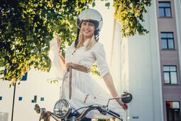 Eine Frau in einem weißen Kleid und Helm auf einem Motorrad in Retro-Stil.
